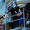 People standing on the deck of a ship near a large light fixture, with colorful flags displayed in the background.