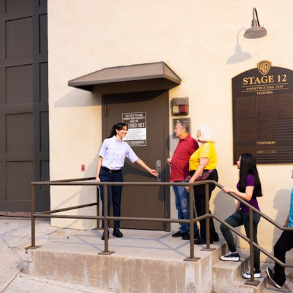 A guide leads a group of visitors into a building labeled Stage 12 at Warner Bros. Studios. A sign lists productions filmed there.