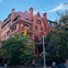 A red brick building with ornate architecture and a steep roof, surrounded by trees, with a streetlight in the foreground.