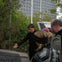A group of people observing a plaque or map outdoors with trees and a large building in the background.