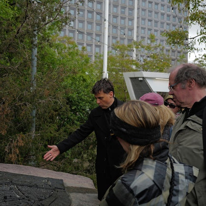 A group of people observing a plaque or map outdoors with trees and a large building in the background.