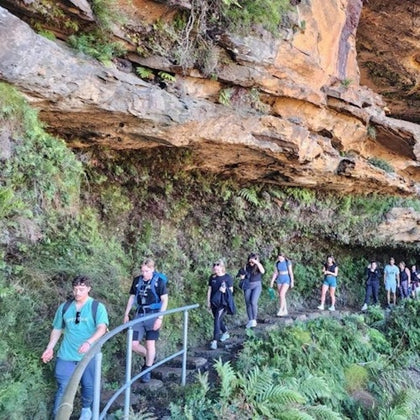 A group of people hiking along a narrow, rocky path lined with ferns and green vegetation under an overhanging rock formation.