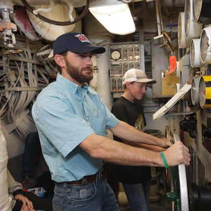 Two men operate machinery in an industrial setting with gauges and wires visible around them.