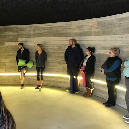 A group of people standing along a curved concrete wall, some holding yoga mats, in a well-lit indoor space.
