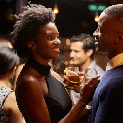 A man and a woman smiling at each other while holding drinks at a social gathering with people in the background.