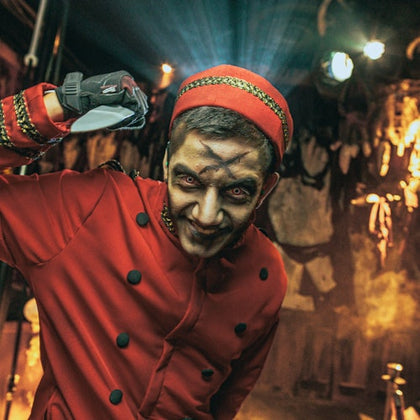 A man in a red uniform and hat with makeup resembling a sinister expression, standing in a dimly lit room with eerie decor.