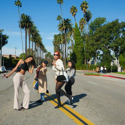 Four people posing playfully on a sunny street lined with tall palm trees, with others visible in the background.