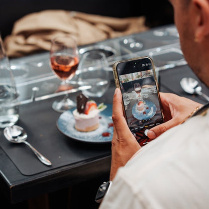 A person takes a photo of a dessert on a blue plate with a smartphone at a table set with glasses, utensils, and a wine glass.