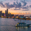 A city skyline at sunset with a ferris wheel and large boat on the water, illuminated buildings in the background.