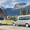A gray van labeled "The Fairy Bus" is parked near a building with mountains in the background under a partly cloudy sky.