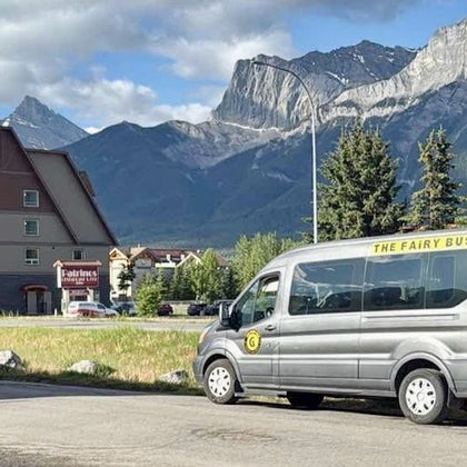 A gray van labeled "The Fairy Bus" is parked near a building with mountains in the background under a partly cloudy sky.