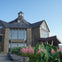 A stone building labeled "Welcome Centre" with large windows, surrounded by greenery and pink flowers under a blue sky.