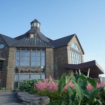 A stone building labeled "Welcome Centre" with large windows, surrounded by greenery and pink flowers under a blue sky.