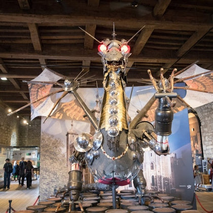 A metallic dragon sculpture with red glowing eyes and wings displayed indoors, with several people observing in the background.