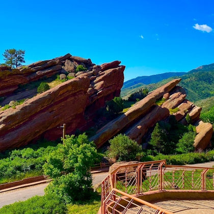 A scenic view of large red rock formations surrounded by greenery under a clear blue sky, with a pathway and railing in the foreground.