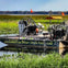 An airboat labeled "Boggy Creek Airboat Rides" is docked on a grassy waterway, equipped with seats, fan, and orange flags.