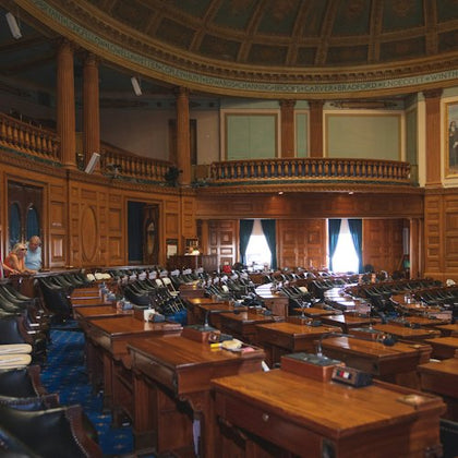 A legislative chamber with empty desks and seating, detailed woodwork, and a domed ceiling. Two people stand at the back.