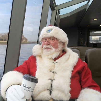 A man dressed as Santa Claus holding a coffee cup on a boat with large windows showing a scenic river view.
