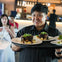 A smiling server holds a tray with plated meals in a restaurant with dining patrons in the background.