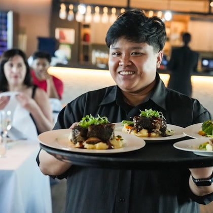 A smiling server holds a tray with plated meals in a restaurant with dining patrons in the background.