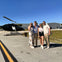 Four people standing in front of a parked black helicopter on an airstrip under a clear blue sky.