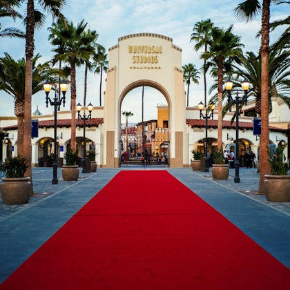 A red carpet leads to the Universal Studios Hollywood entrance arch, surrounded by palm trees and decorative lighting.