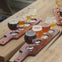 Wooden trays holding small glasses of beer in various shades placed on a wooden table outdoors.