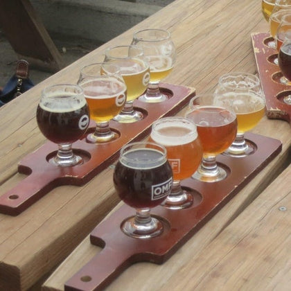 Wooden trays holding small glasses of beer in various shades placed on a wooden table outdoors.