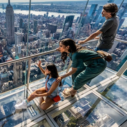 Two people observing a cityscape from a glass observation deck, with a scenic view of buildings and a river in the background.