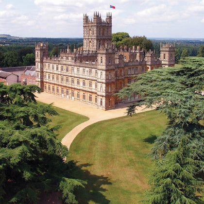 A large, ornate castle with towers and a flag on top, surrounded by green lawns and trees under a partly cloudy sky.