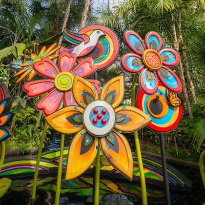 Colorful artistic sculptures of flowers and a bird surrounded by lush tropical plants in a garden setting.
