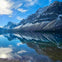 Snow-capped mountains reflected in a clear, still lake under a bright blue sky with scattered clouds.