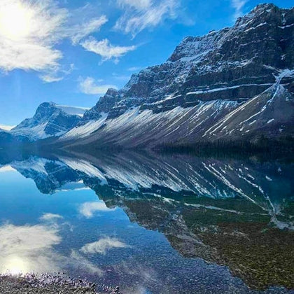 Snow-capped mountains reflected in a clear, still lake under a bright blue sky with scattered clouds.