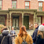 A group of people stand in front of a historic brick building with green doors and large windows, chatting and smiling.