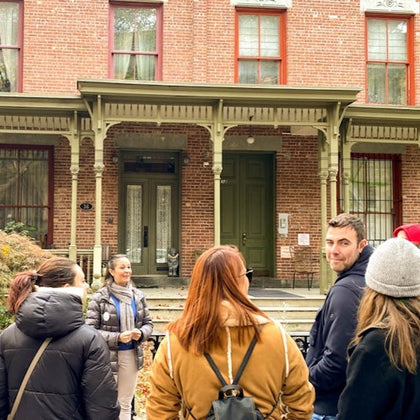 A group of people stand in front of a historic brick building with green doors and large windows, chatting and smiling.
