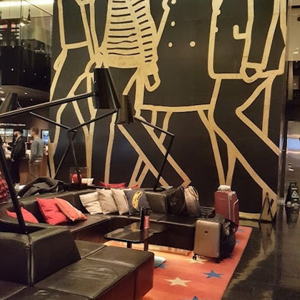 A modern lobby with a large mural of walking figures, leather seating, cushions, floor lamps, and suitcases on the floor.