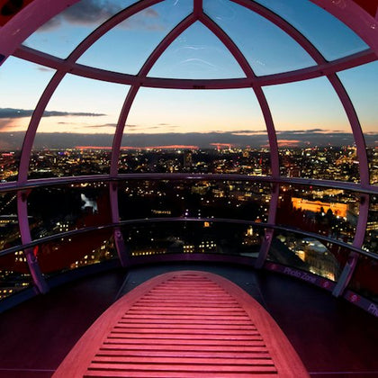 Interior of a Ferris wheel capsule overlooking a cityscape at dusk, with illuminated buildings and a red bench in the foreground.