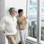 Two men stand indoors by a large window overlooking a cityscape with tall buildings, water, and a partly cloudy sky.