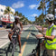 Two people on bicycles converse on a palm-lined urban street with a designated bike lane. Buildings and parked cars are in the background.