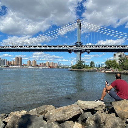 Man sits on rocks by a river, looking at a suspension bridge with city skyline and blue sky with clouds in the background.
