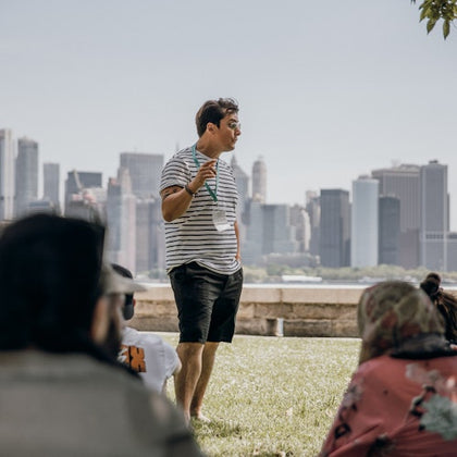 A man stands outside speaking to a small group, with a city skyline in the background.