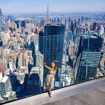 A person taking a selfie on a rooftop with a view of a city skyline, including the Empire State Building in the distance.