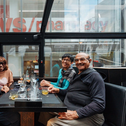 Three people sit smiling at a table with drinks and empty glasses. The background shows a glass wall with the word "pharmacy."