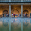A group of people stand by the edge of a steaming water pool, surrounded by ancient stone columns and walls.