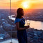 A woman in a blue dress holds a glass of wine while looking out at a cityscape and river during sunset from a tall building.