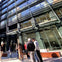 Several people stand talking in front of a modern glass building on a sunny day.