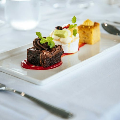 A dessert platter featuring a brownie with chocolate garnish, a fluffy cake with fruit and cream, and a yellow cake with icing on a white tablecloth.