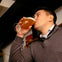 A person in a gray sweater drinks a glass of beer in an indoor setting.