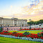 A grand building with a large courtyard, colorful flowerbeds, and people walking, set against a sunset sky.