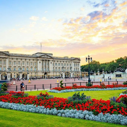 A grand building with a large courtyard, colorful flowerbeds, and people walking, set against a sunset sky.